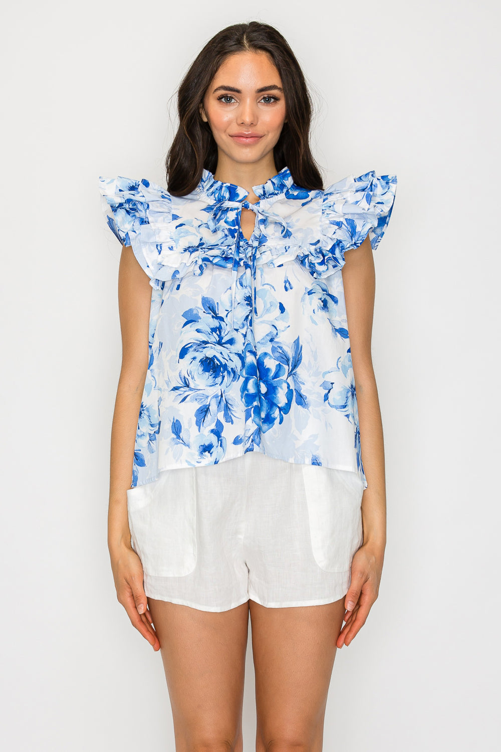 Model wearing a blue floral ruffle-sleeve blouse with tie keyhole neckline and flared silhouette, styled with white linen shorts.