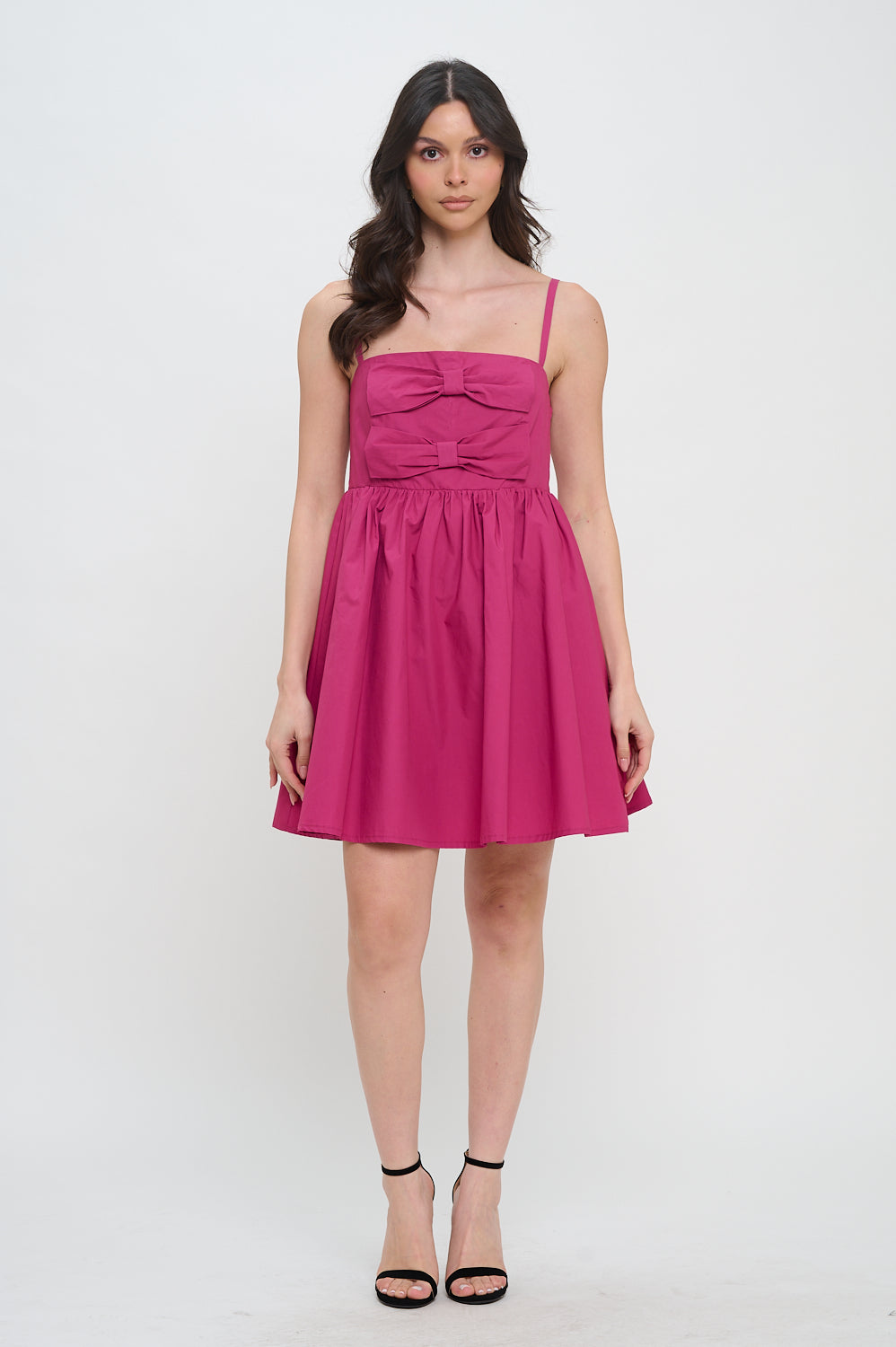 Model wearing a deep pink mini dress with spaghetti straps, bow details on the bodice, and a gathered skirt.