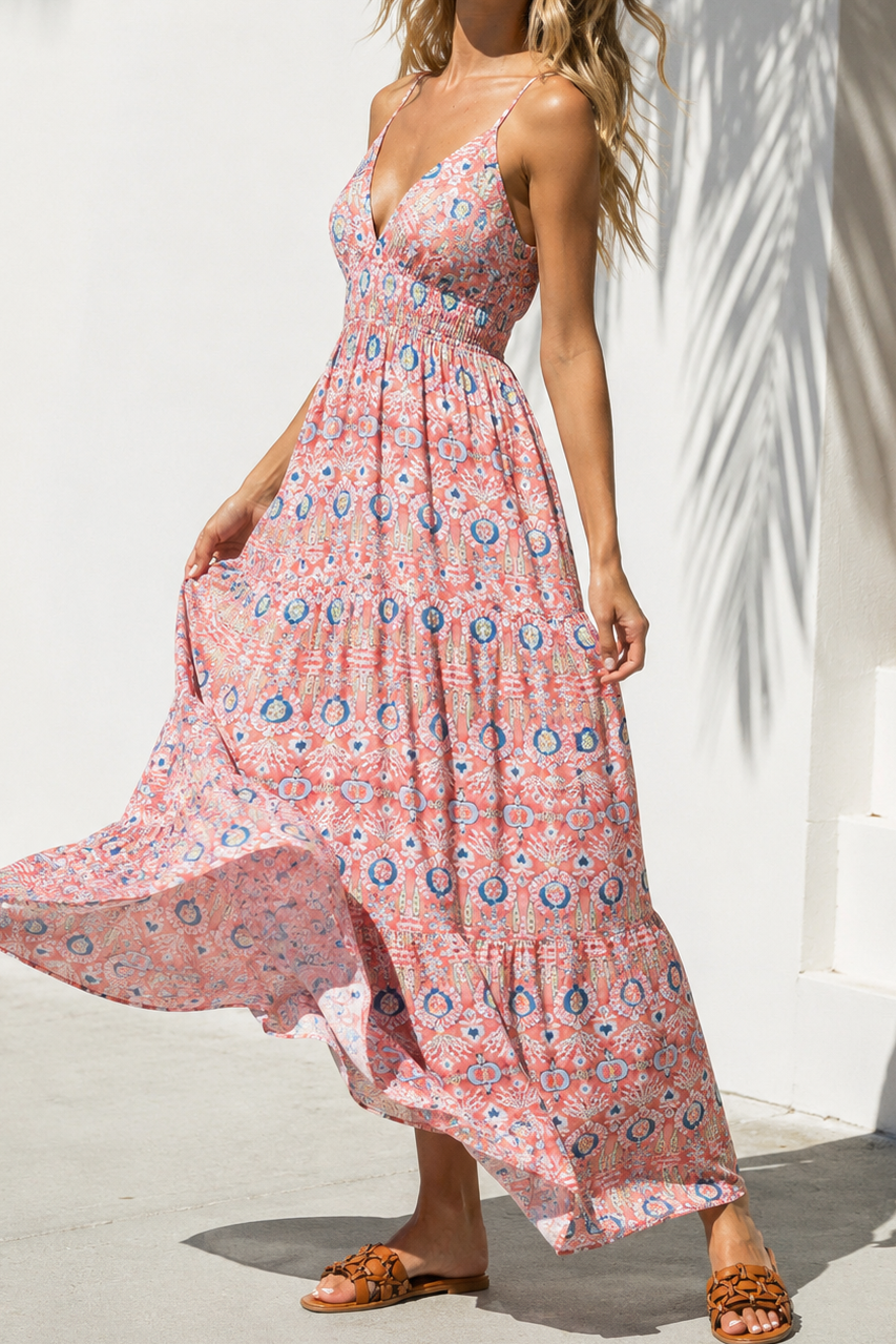Front view of a blonde model standing near a white wall under a palm shadow, wearing the Sunset Tapestry Maxi Dress, a rose pink and blue tiered maxi dress with a plunge V-neck and a smocked empire waist.