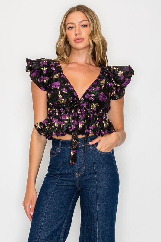 Model wearing the Nyx Petals Adelina Top, a black and purple metallic floral top with a V-neck, ruffle sleeves, and a peplum hem.