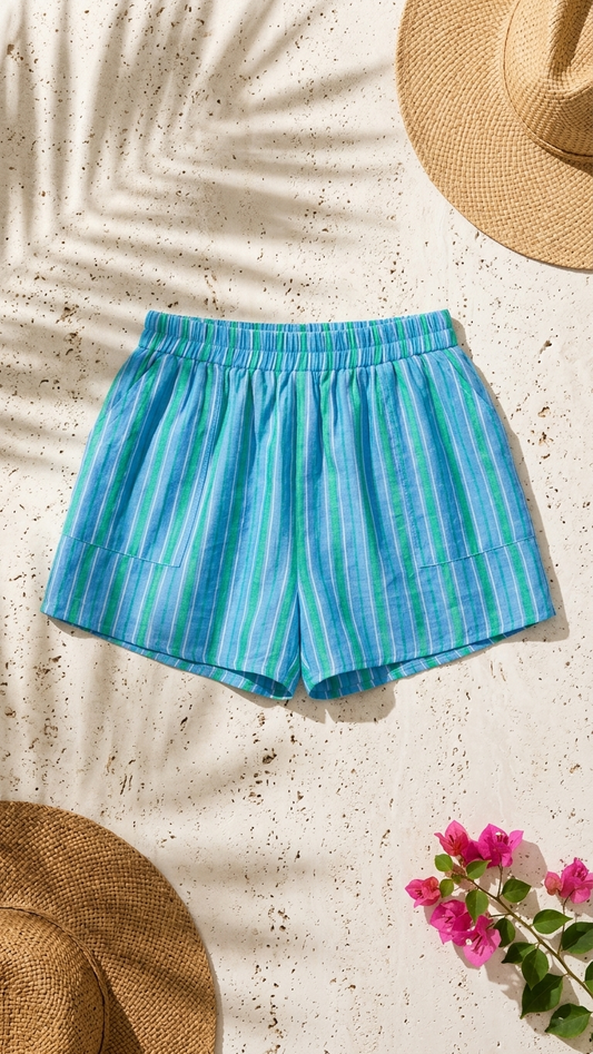 Flat lay of the Coastal Drift Shorts, azure blue and green striped cotton shorts with an elastic waist on a stone background with palm shadows and a straw hat.