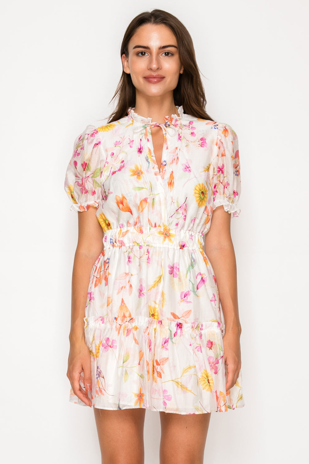 Model wearing the Flora of Eos Short Dress, a white mini dress with a vibrant pink and orange floral print, featuring a ruffled neck, tie detail, and smocked waist.