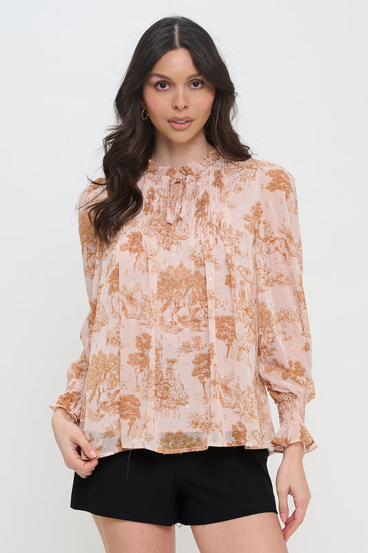 Woman wearing a chiffon long sleeve blouse with a soft pink and copper pastoral print, metallic accents, tie neck, and smocked cuffs.