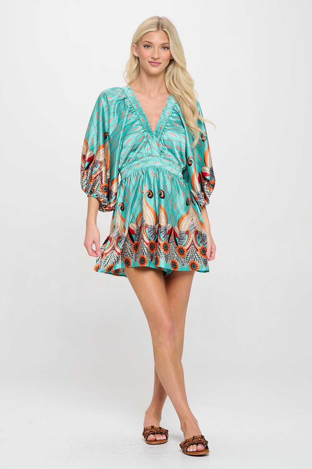 Model wearing a teal printed romper with dolman sleeves, a ruched V-neckline, and feather-patterned border hem.