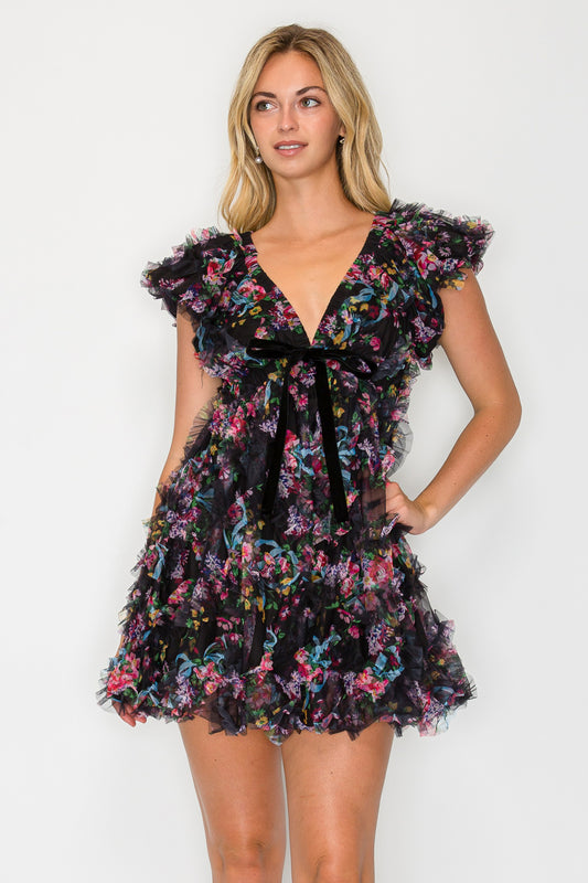 Model wearing the Whisper Bloom Flutter Sleeve Mini Dress, a black floral tulle dress with a V-neck, velvet bow, and dramatic flutter sleeves.