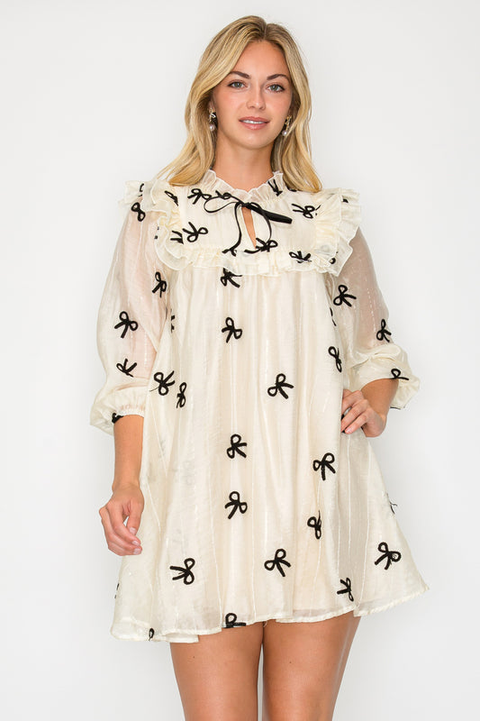 Model wearing the Organza Bow Long Sleeve Mini Dress, a cream-colored dress with a large ruffle collar, bishop sleeves, and black velvet bow details.