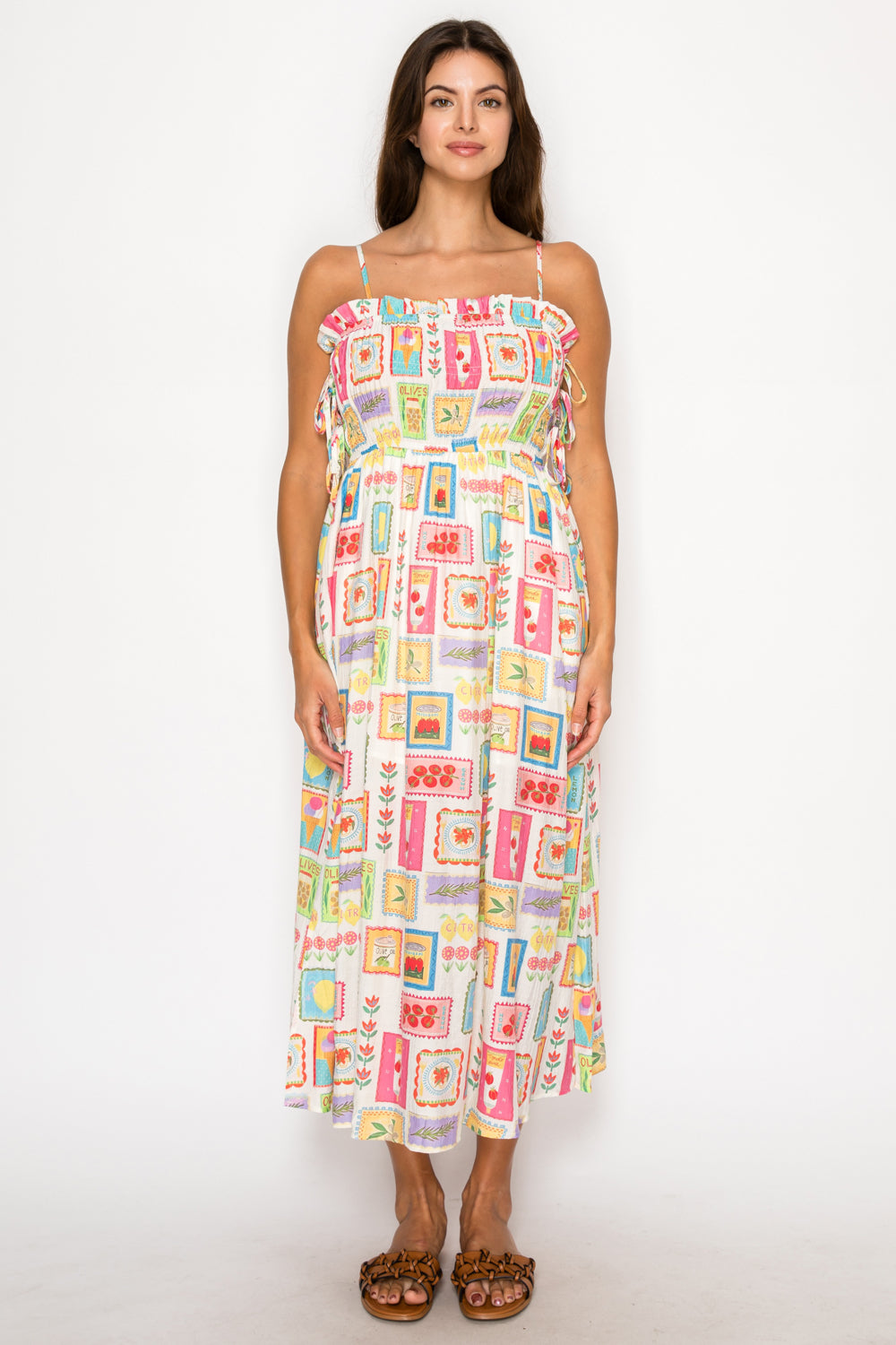 Model wearing the Citrus Kefi Midi Dress, a white midi dress with a colorful postage-stamp style print of citrus and olives, featuring a smocked bodice and ruffles.