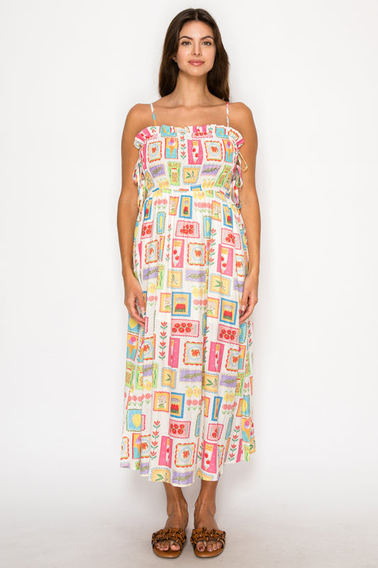 Model wearing the Citrus Kefi Midi Dress, a white midi dress with a colorful postage-stamp style print of citrus and olives, featuring a smocked bodice and ruffles.