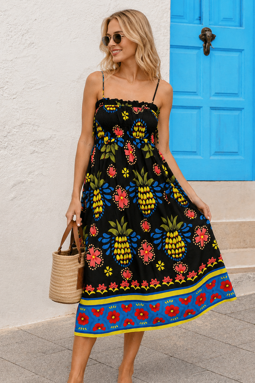 Front view of a blonde model standing next to a white wall with a bright blue door, wearing the Ionian Charm Midi Dress, a black smocked midi dress with a colorful yellow pineapple and red floral folk print.
