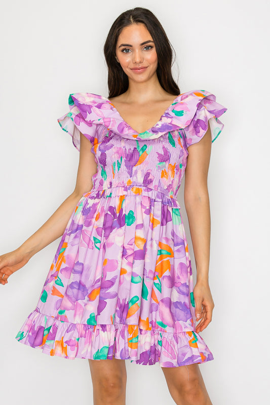 Model wearing a floral mini dress with smocked bodice, ruffle neckline, and flouncy hem in a lilac and multicolor print.