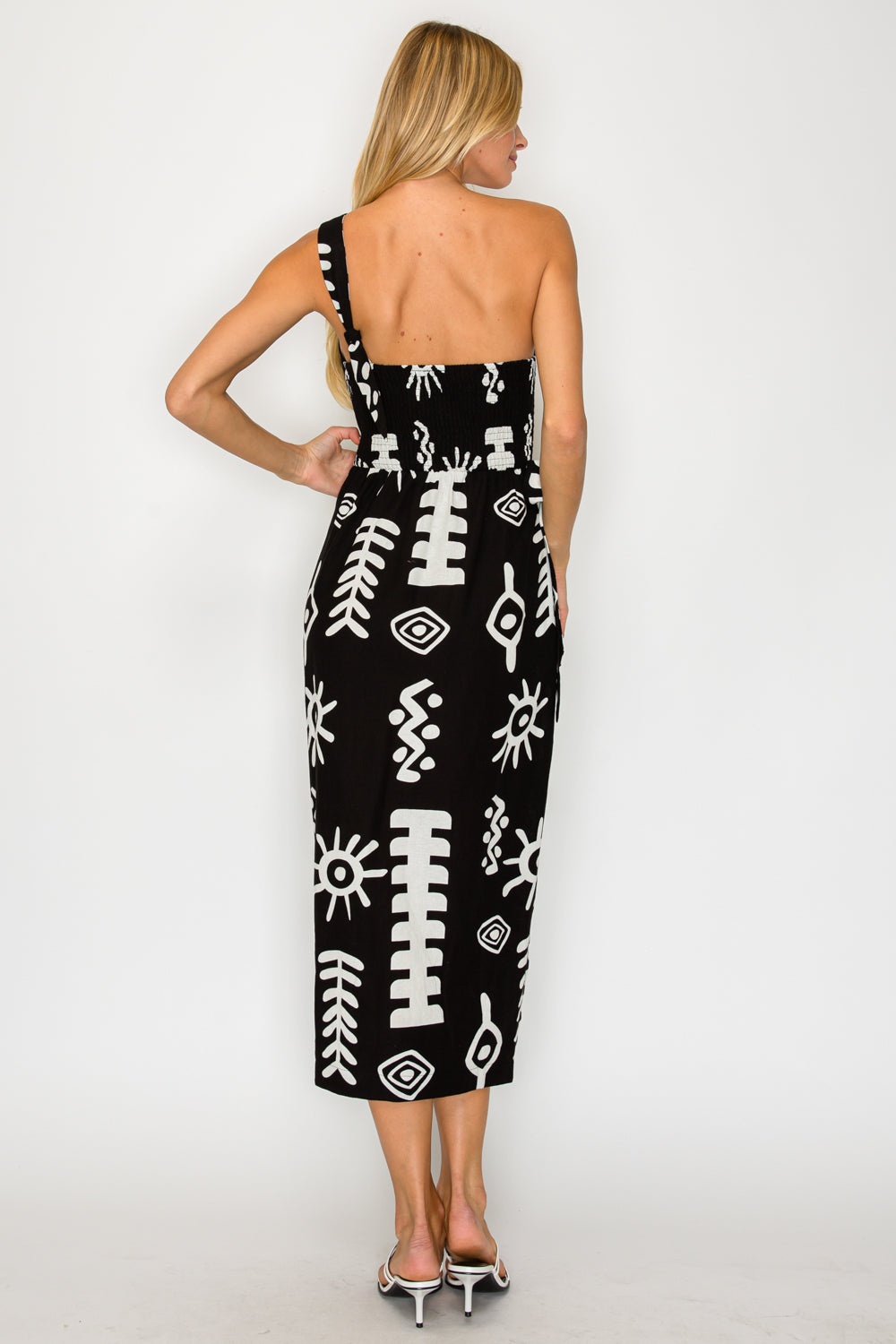 Front-side view of a model standing near a whitewashed sunlit wall, wearing the Paros Geometry One Shoulder Dress, a black midi dress with a white graphic block-print pattern.