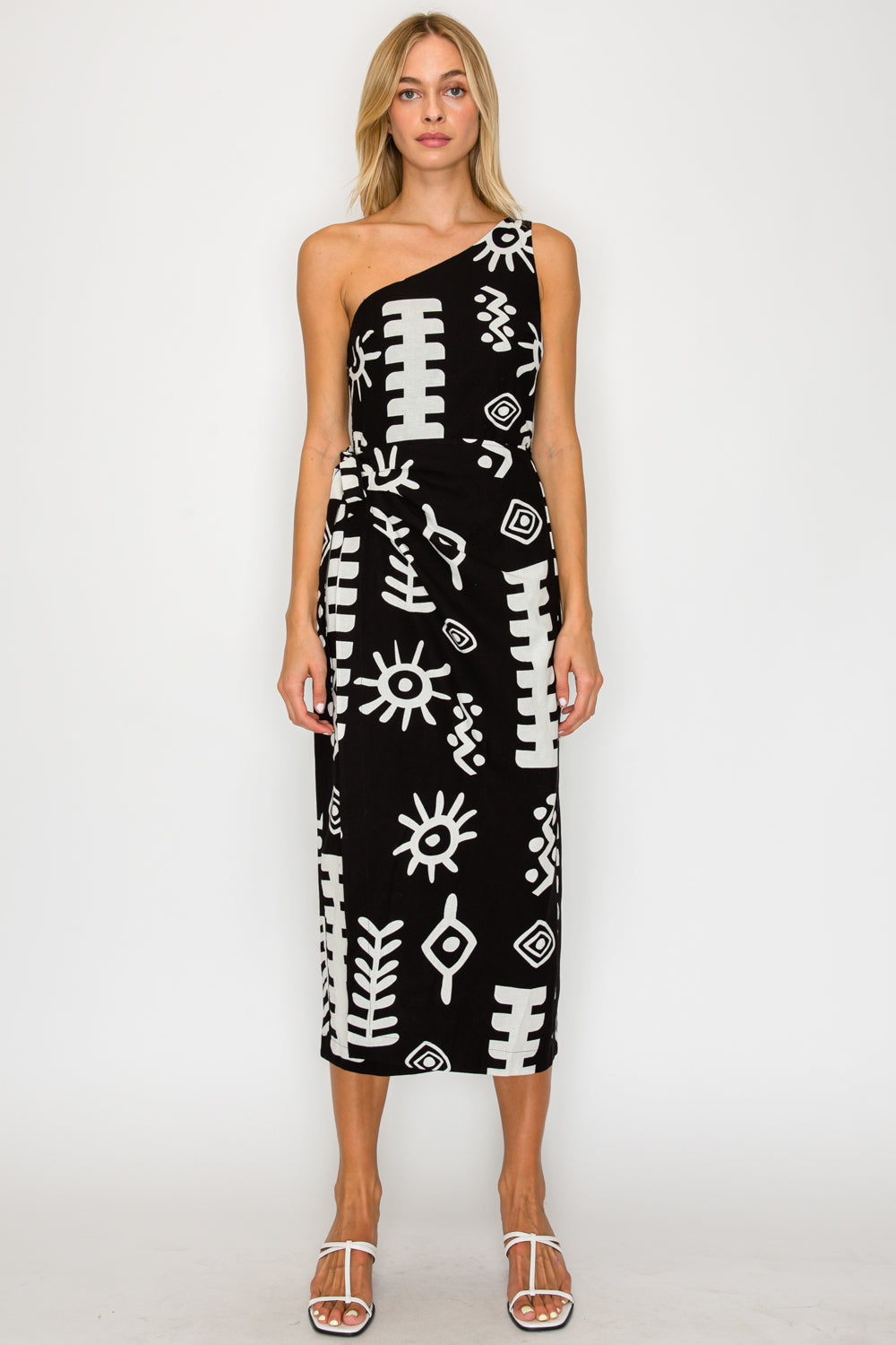 Front-side view of a model standing near a whitewashed sunlit wall, wearing the Paros Geometry One Shoulder Dress, a black midi dress with a white graphic block-print pattern.