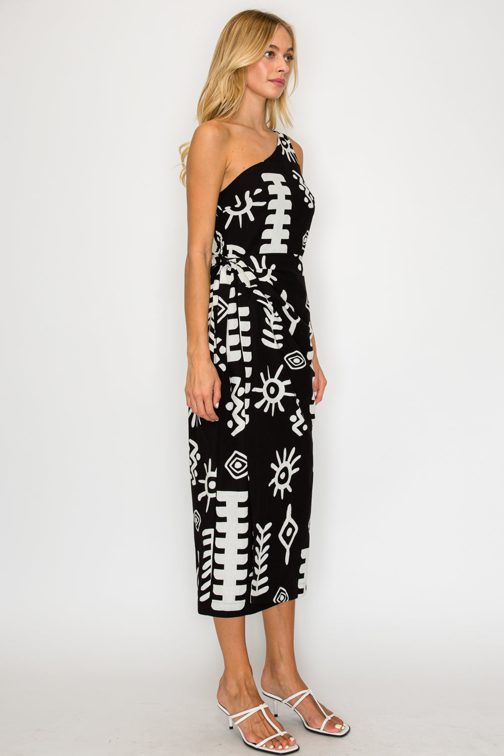 Front-side view of a model standing near a whitewashed sunlit wall, wearing the Paros Geometry One Shoulder Dress, a black midi dress with a white graphic block-print pattern.