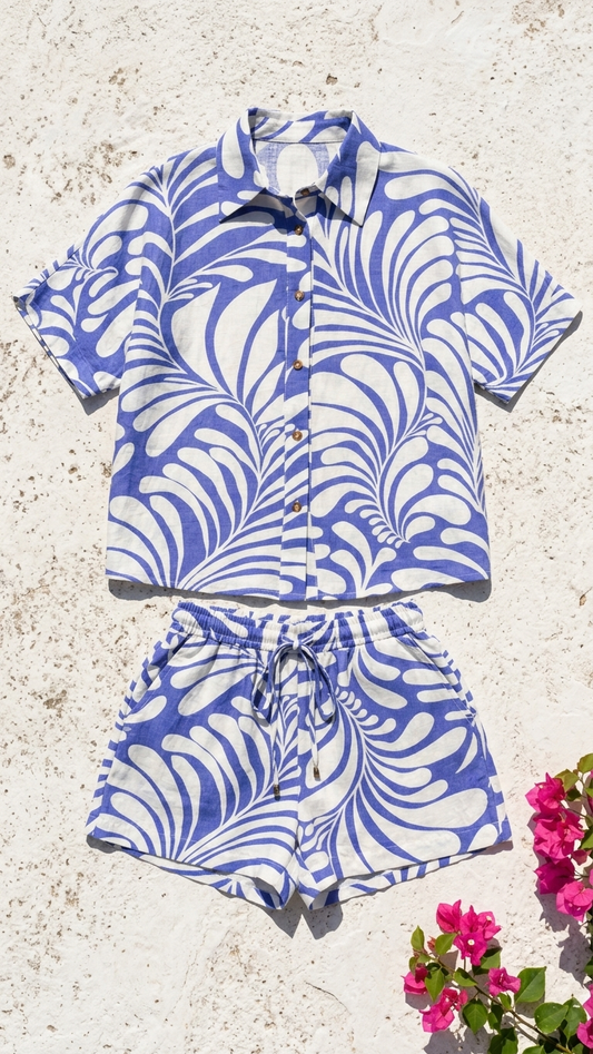 Flat lay of the Cycladic Palm Set, a blue and white abstract palm print two-piece shirt and shorts set on a white stone background with bougainvillea.