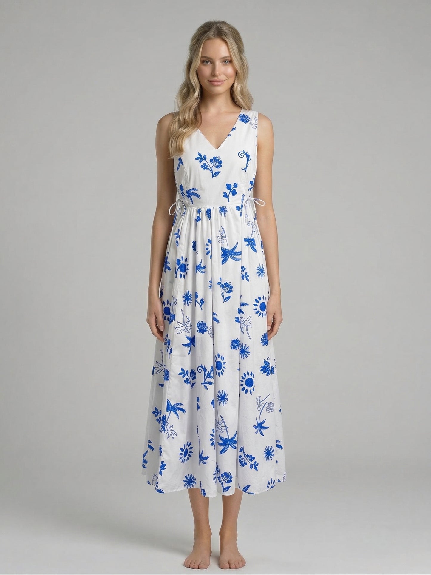 Model wearing the Island Azure Maxi Dress, a white floor-length dress with a vibrant blue tropical and floral print, featuring a V-neckline and side cut-outs with ties.