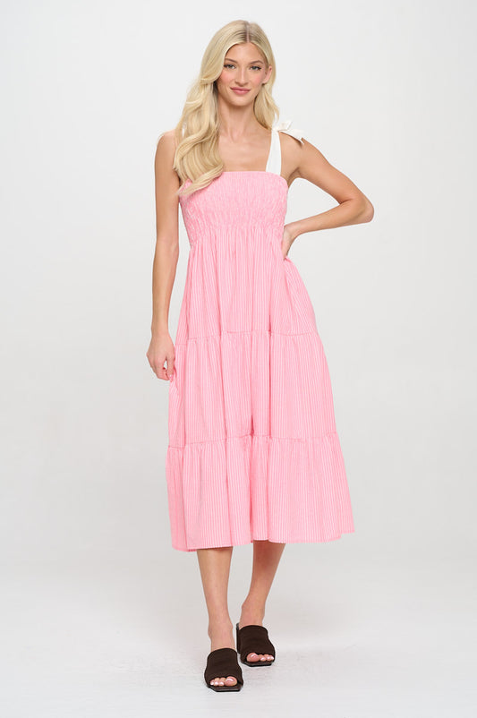 Model wearing a pink and white striped midi dress with smocked bodice and tiered skirt, styled with wide self-tie shoulder straps.