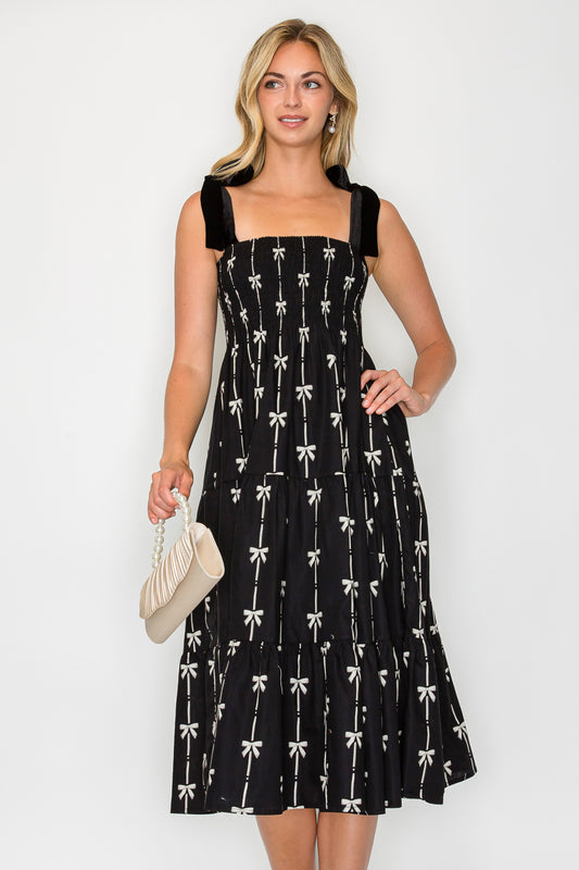 Model wearing the Holiday Bow Midi Dress, a black midi dress with a bow print, smocked bodice, and velvet tie-shoulders.