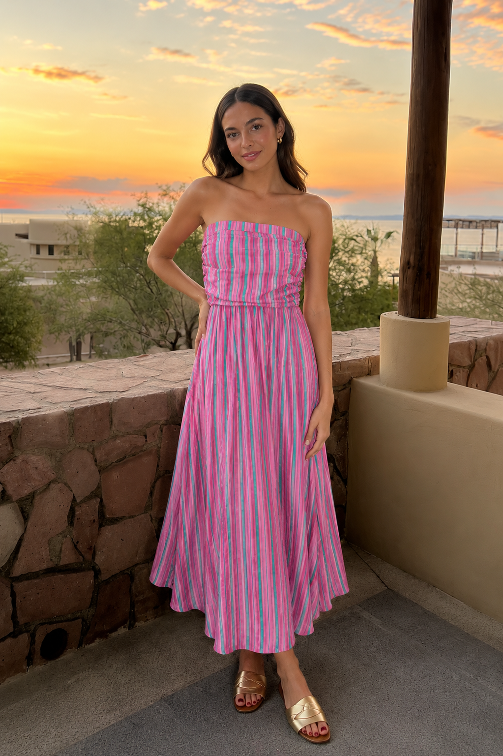 Woman in a pink striped dress standing on a balcony with a sunset view.
