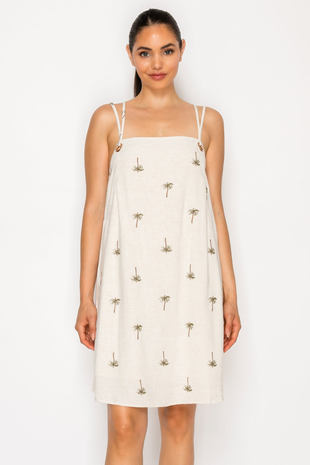 Model wearing the Drifting Palms Mini Dress, a cream-colored shift dress with embroidered palm trees and button-detail straps.