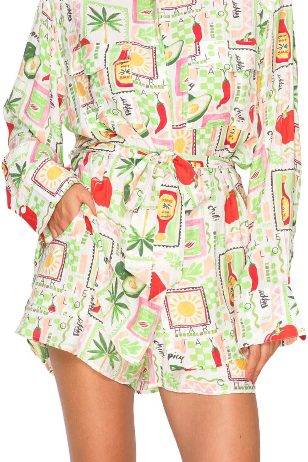 Model wearing the Pepper Parea Shorts, a white pair of shorts with a colorful chili pepper and avocado graphic print, featuring a matching tie-belt.