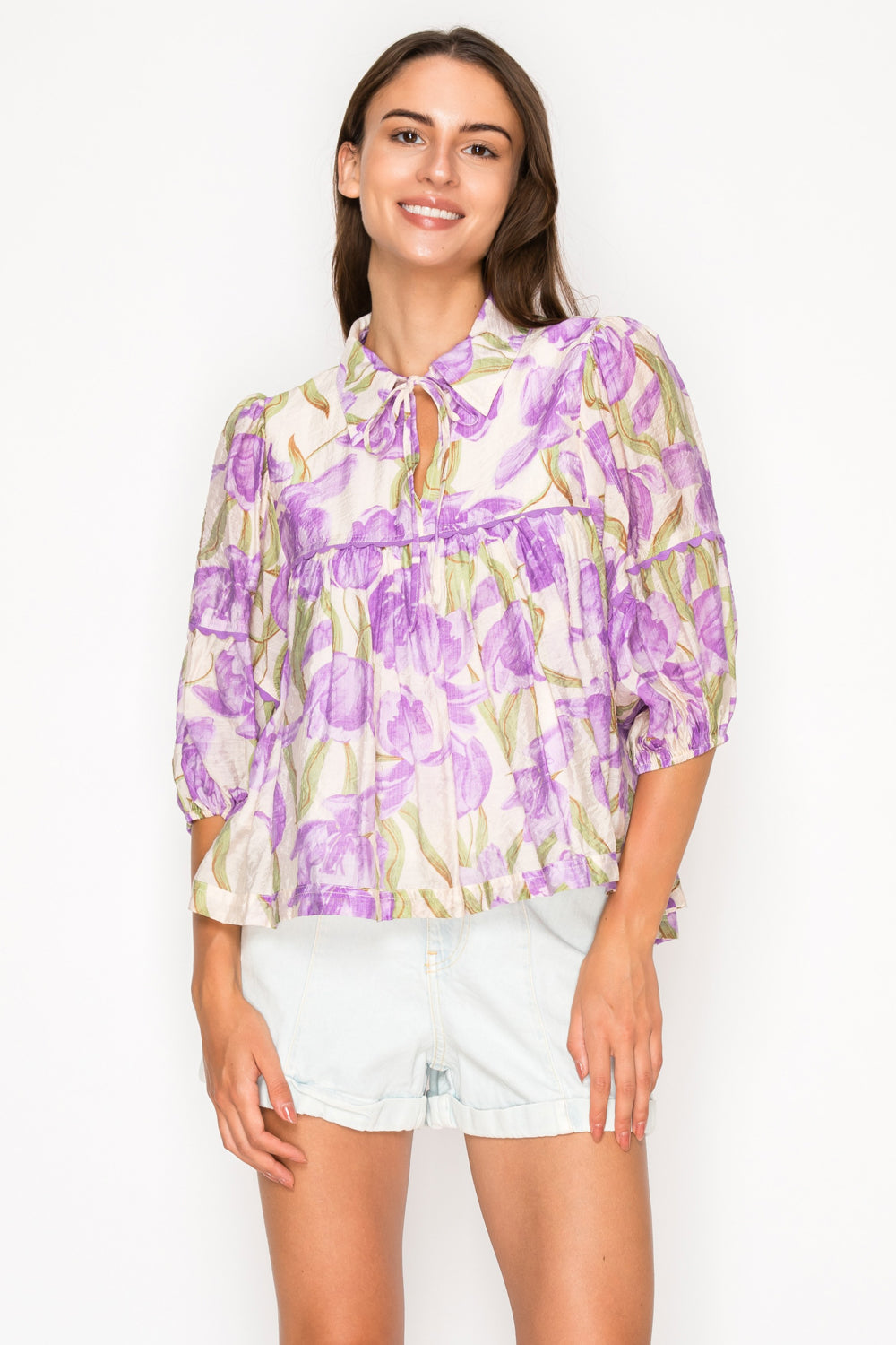 Model wearing the Aphrodites Garden Blouse, a purple floral print top with a tie-neck, puff sleeves, and a tiered babydoll silhouette.