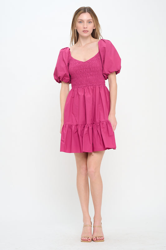 Model wearing a deep pink mini dress with puff sleeves, a smocked bodice, and a tiered ruffle hem.