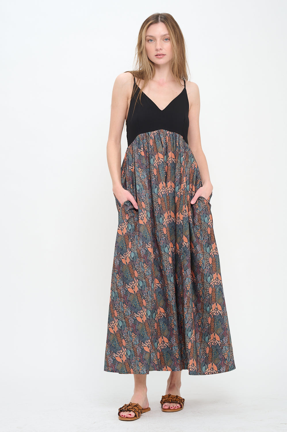 Model wearing a sleeveless V-neck maxi dress with a black fitted bodice and printed autumnal forest skirt, styled with flat woven sandals.