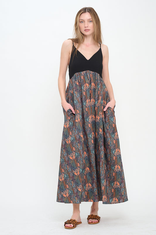 Model wearing a sleeveless V-neck maxi dress with a black fitted bodice and printed autumnal forest skirt, styled with flat woven sandals.