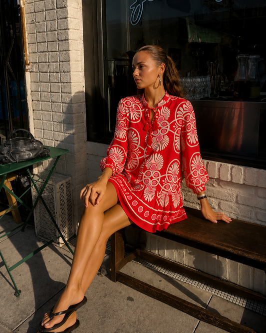 Side-front view of a model sitting on a wooden bench outside, wearing the Festival Medallion Short Dress, a red mini dress with a white and pink fan medallion print, long balloon sleeves, and a tiered ruffle hem.
