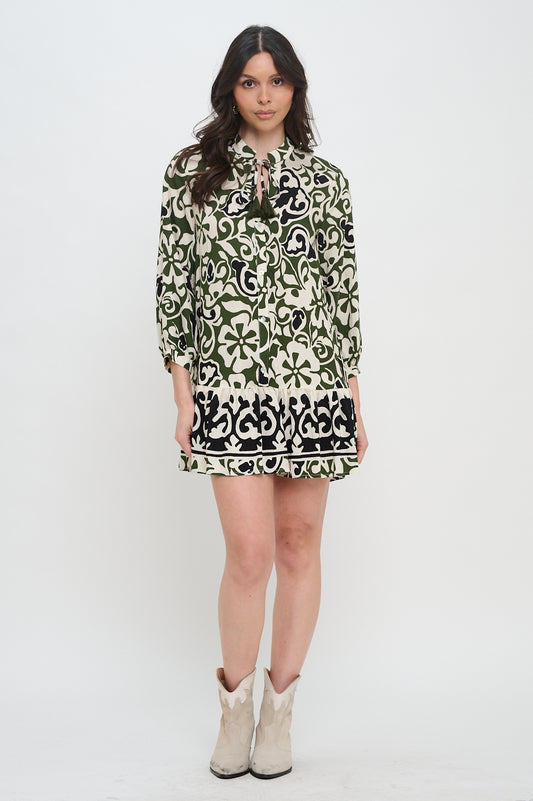 Model wearing olive scroll print long sleeve mini dress with button front and tassel tie neckline
