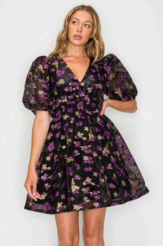 Model wearing the Nyx Petals Short Sleeve Dress, a black and purple metallic floral mini dress with a V-neck, puff sleeves, and a smocked waist.