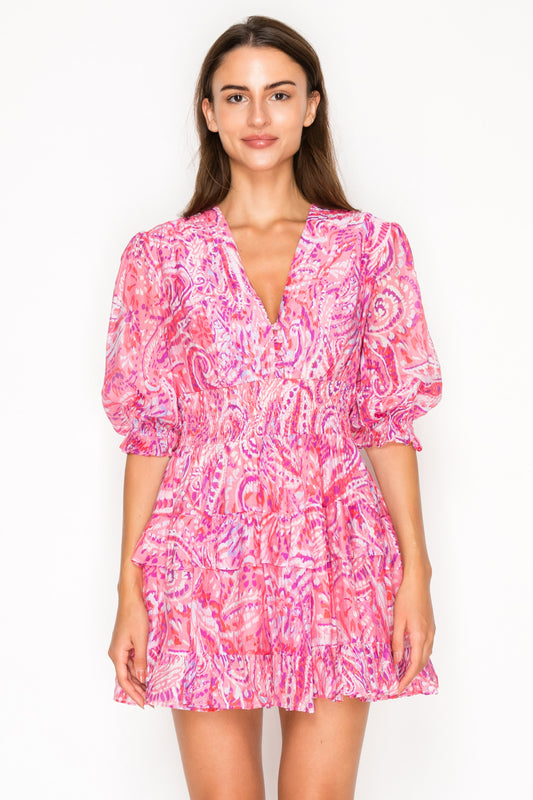 Model wearing the Rosey Echoes Short Dress, a vibrant pink and magenta paisley mini dress with a V-neck, puff sleeves, and a smocked waist.