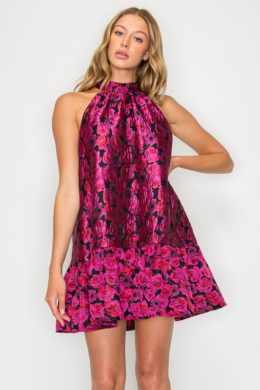 Model wearing the Heras Garland Halter Dress, a pink and black metallic floral mini dress with a high halter neck and a tiered ruffle hem.