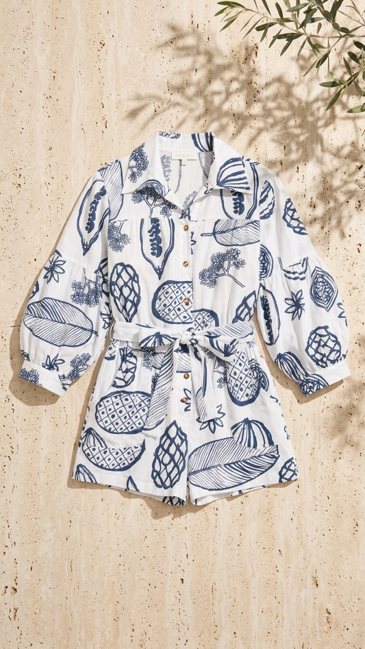 Flat lay of the Fruit Delight Romper, a white linen-blend romper featuring a blue fruit illustration print, long lantern sleeves, and a button-front closure on a stone background.