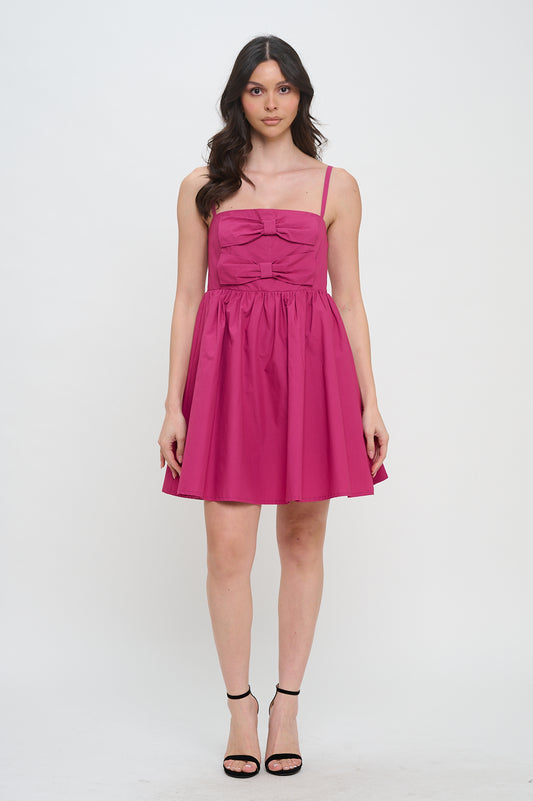 Model wearing a deep pink mini dress with spaghetti straps, bow details on the bodice, and a gathered skirt.