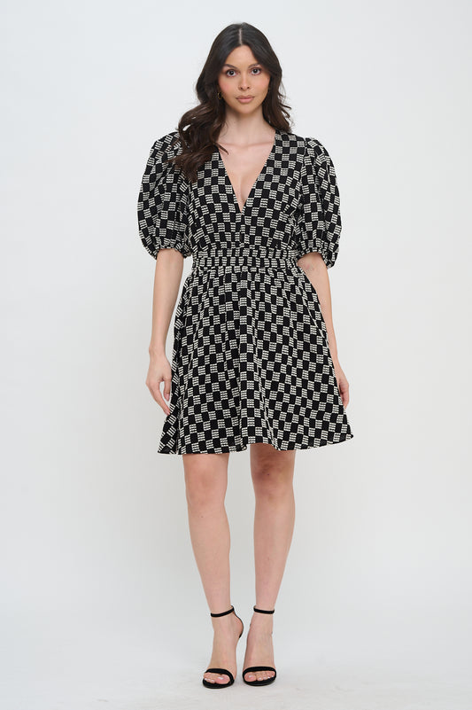 Model wearing the Midnight Weave Short Sleeve Dress from Meet Me in Santorini, a black and white checkerboard mini dress with a V-neck and puff sleeves.