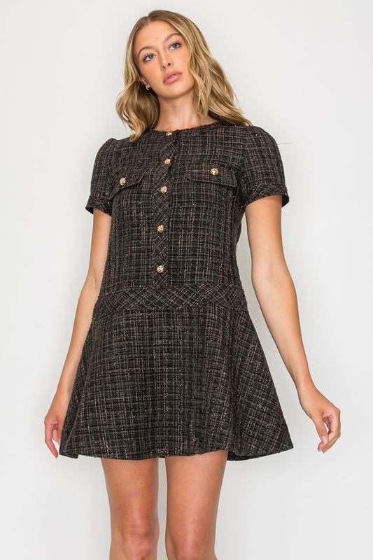 Model wearing the Ebony Tweed Short Sleeve Dress, a black tweed mini dress with gold buttons, chest pockets, and a flared drop-waist skirt.