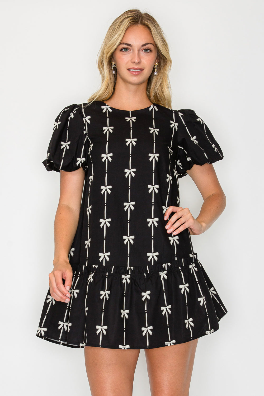 Model wearing the Holiday Bow Mini Dress, a black dress with a cream bow print, puff sleeves, and a drop-waist ruffle hem.