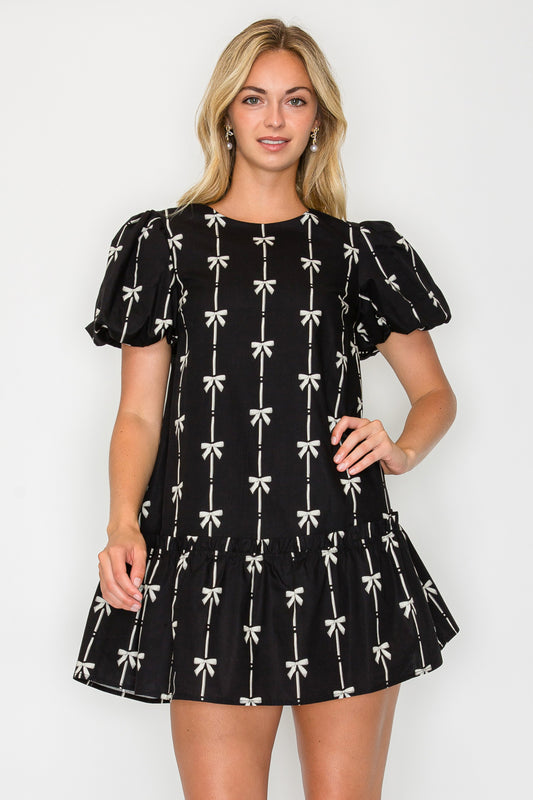 Model wearing the Holiday Bow Mini Dress, a black dress with a cream bow print, puff sleeves, and a drop-waist ruffle hem.