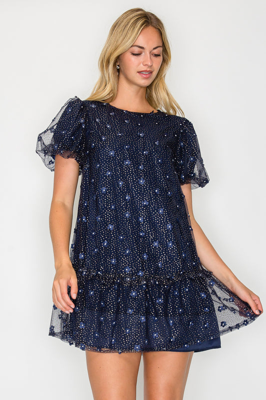 Model wearing the Holiday Daisy Puff Sleeve Mini Dress, a navy tulle dress with embroidered daisies, puff sleeves, and a drop-waist ruffle hem.