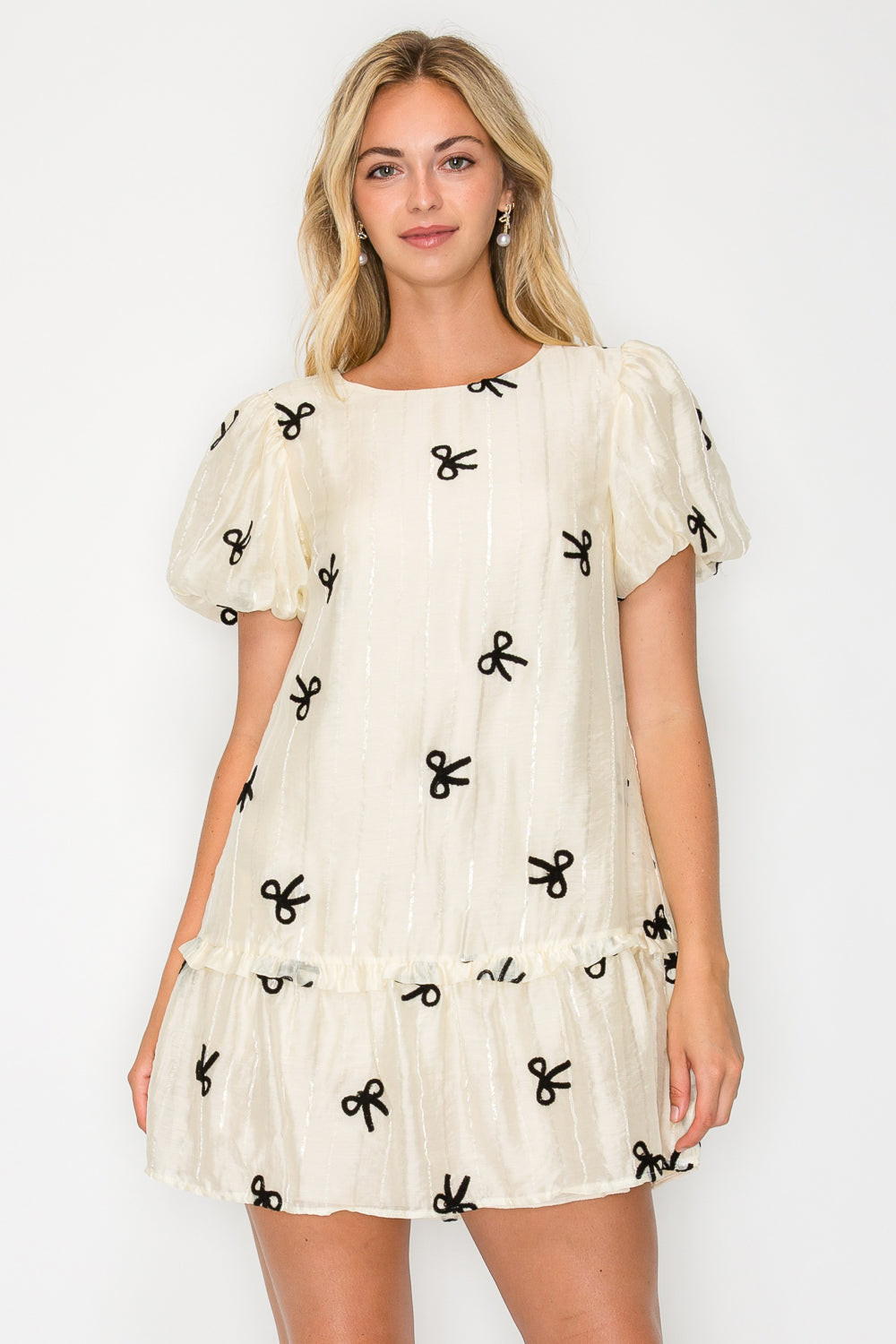 Model wearing the Organza Bow Puff Sleeve Mini Dress, a cream-colored dress with black velvet bow details, puff sleeves, and a drop-waist ruffle hem.