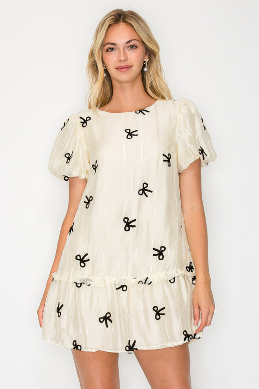 Model wearing the Organza Bow Puff Sleeve Mini Dress, a cream-colored dress with black velvet bow details, puff sleeves, and a drop-waist ruffle hem.