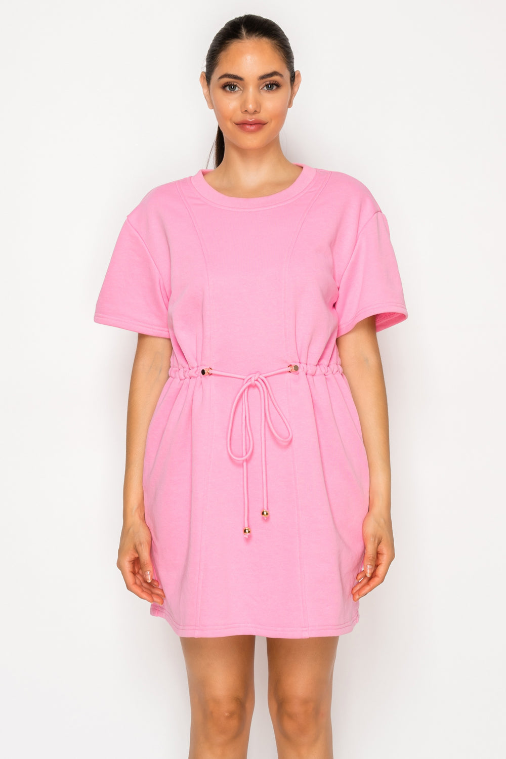 Model wearing the Pink French Terry Short Dress, a solid pink mini dress with short sleeves, a crew neck, and a drawstring cinched waist with gold hardware.