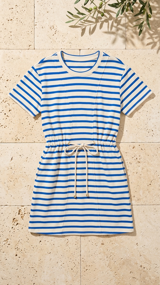 Flat lay of the Sapphire Waves Dress, a blue and ivory horizontal striped t-shirt dress featuring a drawstring waist and gold-tipped ties on a stone background with olive branches.