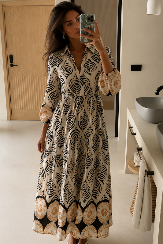 Mirror selfie of a woman wearing the Midnight Shell Collared Maxi Dress, a black and cream shell-printed tiered maxi dress with balloon sleeves, a pointed collar, and a terracotta medallion border hem.