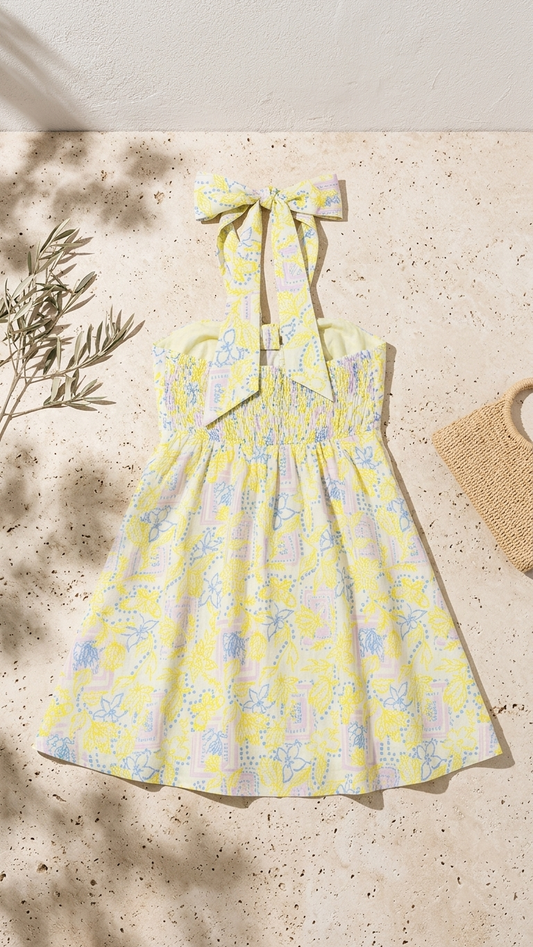 Flat lay of the Lemon Breeze Halter Dress, a yellow and lavender floral print mini dress featuring a large halter neck bow and a smocked back, shown next to an olive branch and straw bag.