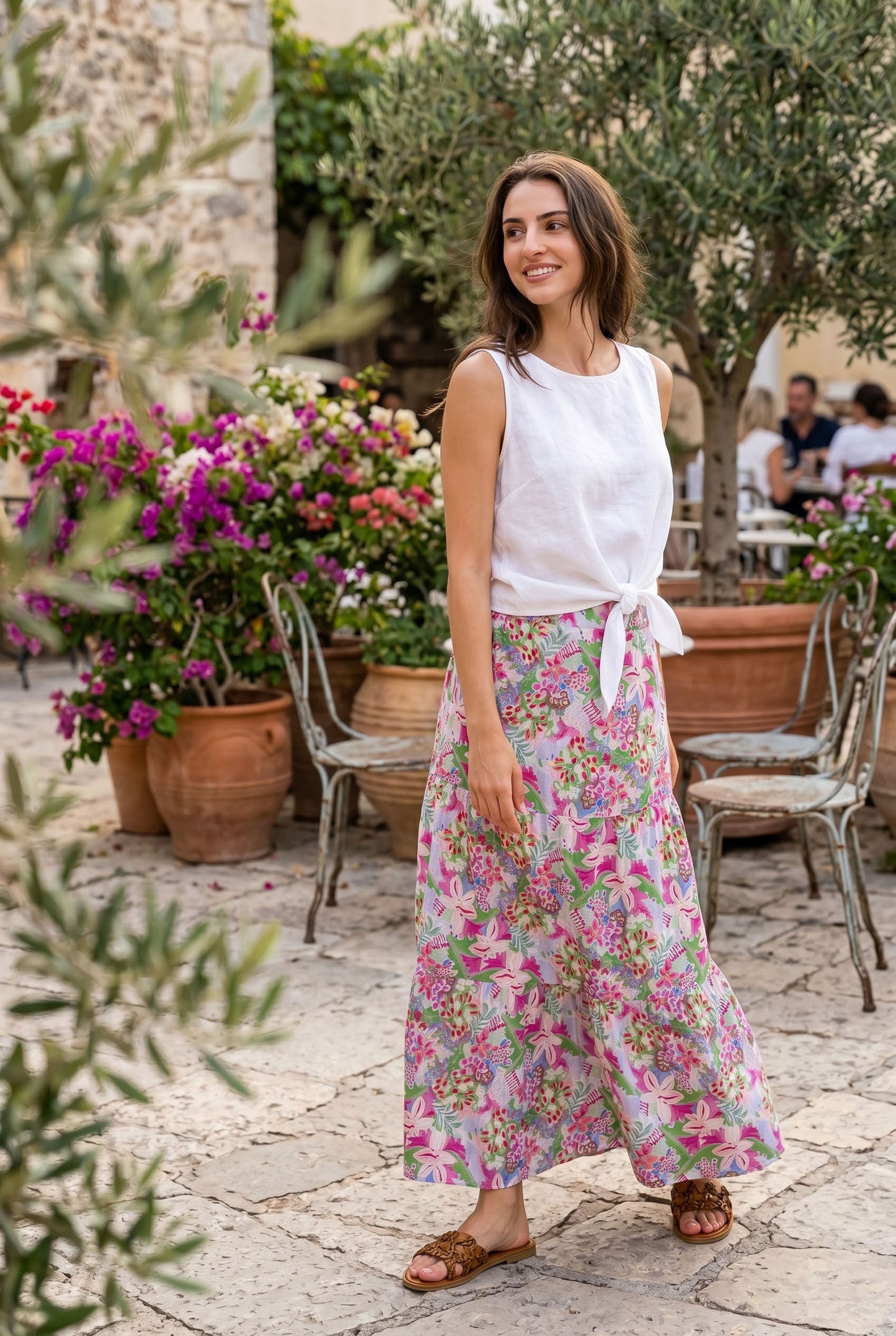 Woman in a white top and floral skirt standing in an outdoor setting with flowers and people in the background.