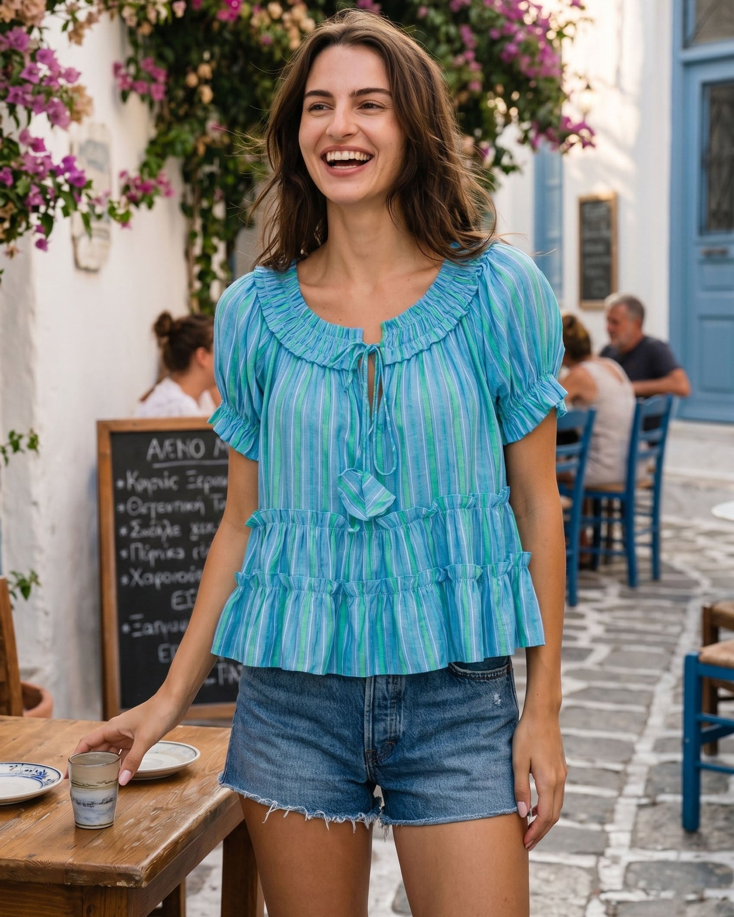 Woman in a blue striped blouse and denim shorts standing in a picturesque street with flowers and a cafe.